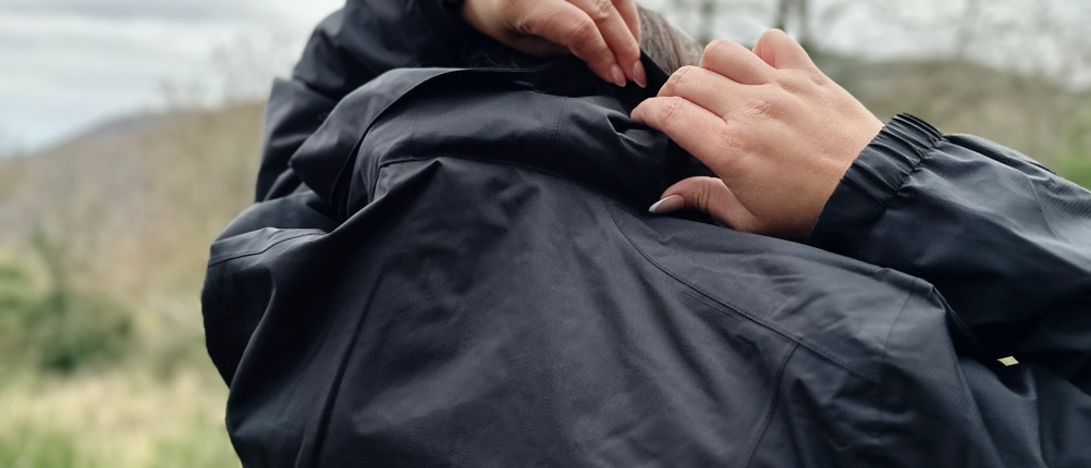 Die Frau, die die schwarze Vaude Regenjacke trägt, packt die zuvor im Kragen verstaut gewesene Kapuze aus.