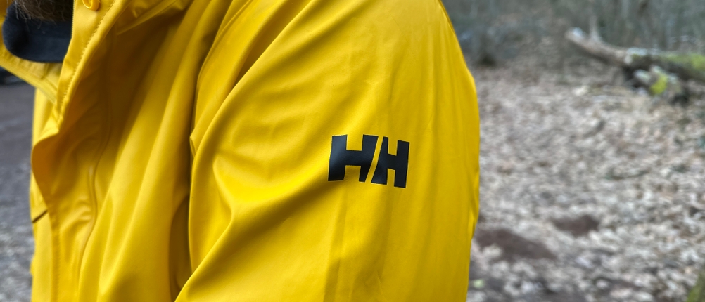 Nahaufnahme des HH-Logos auf dem Ärmel der Helly Hansen Moss Regenjacke. Die schwarze Schrift hebt sich vom gelben Stoff ab.