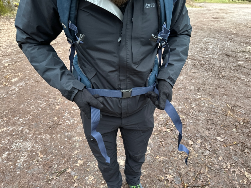 Ein Mann trägt die Marmot Minimalist Regenjacke und einen Rucksack, während er die Brusttasche nutzt. Die Tasche ist trotz der Schultergurte gut erreichbar.