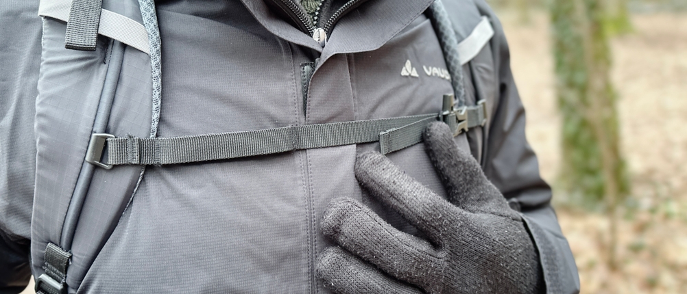 Ein Mann trägt die Vaude Escape Light Regenjacke mit einem Rucksack. Der Brustgurt des Rucksacks liegt über der Jacke.