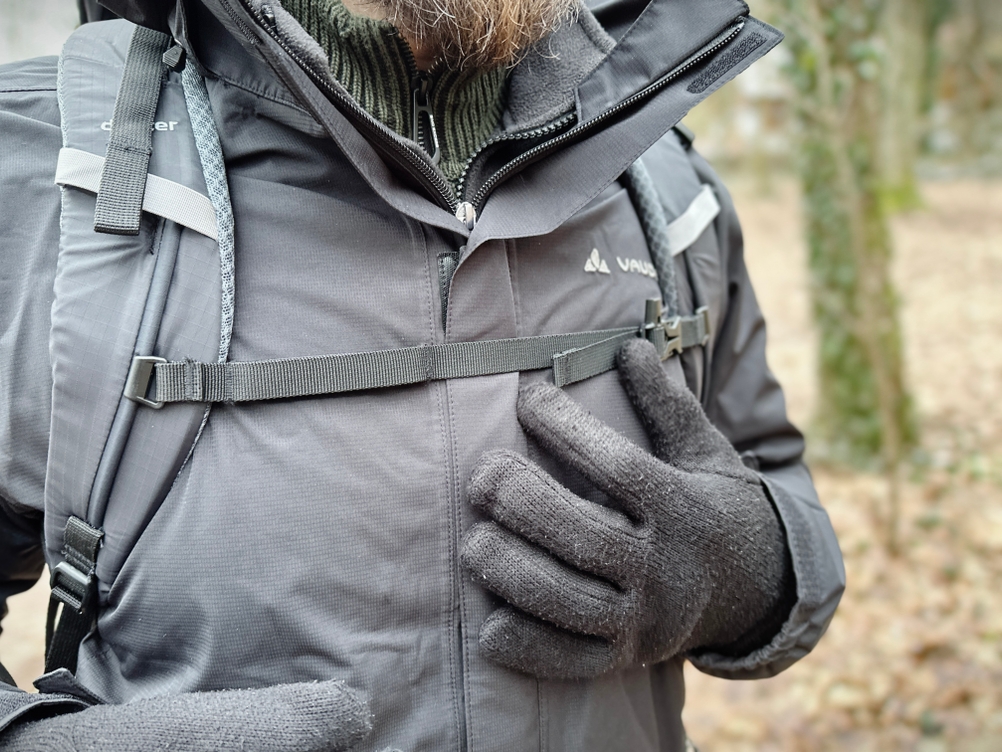 Ein Mann trägt die Vaude Escape Light Regenjacke mit einem Rucksack. Der Brustgurt des Rucksacks liegt über der Jacke.
