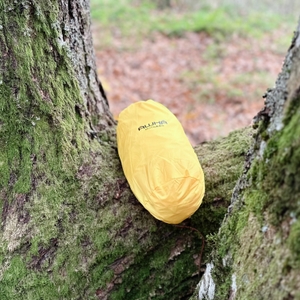 Der gelbe Regenponcho AWHA im Beutel liegt auf einem Baumrahmen.