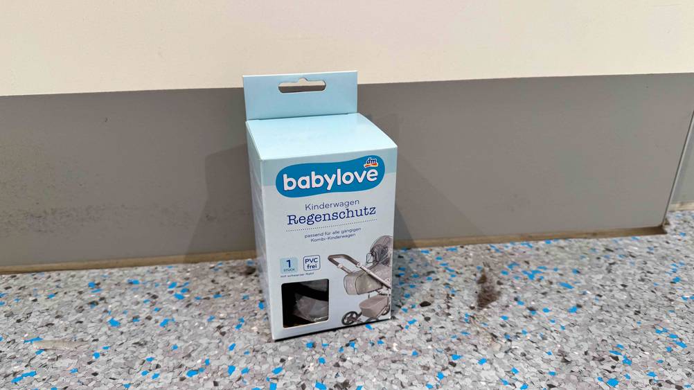 Der Regenschutz Kinderwagen im Test in der Originalverpackung vor einer Wand auf dem Boden.