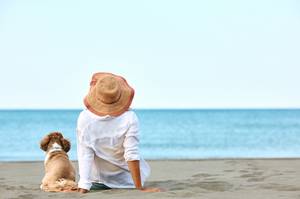 Reiserücktrittsversicherungen getestet: Eine Frau sitzt mit ihrem Hund an einem Strand und blickt Richtung Meer.
