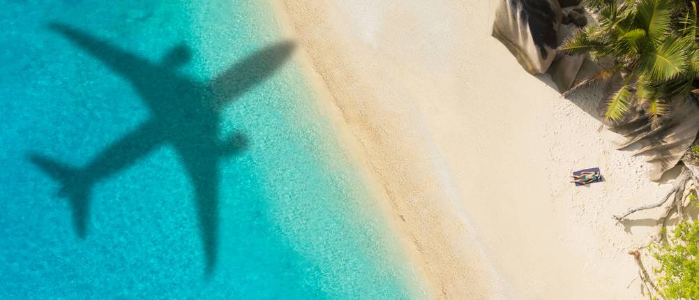 Reiserücktrittsversicherung-Test: Auf einer Luftaufnahme eines Strands ist der Schatten eines Flugzeugs zu sehen.
