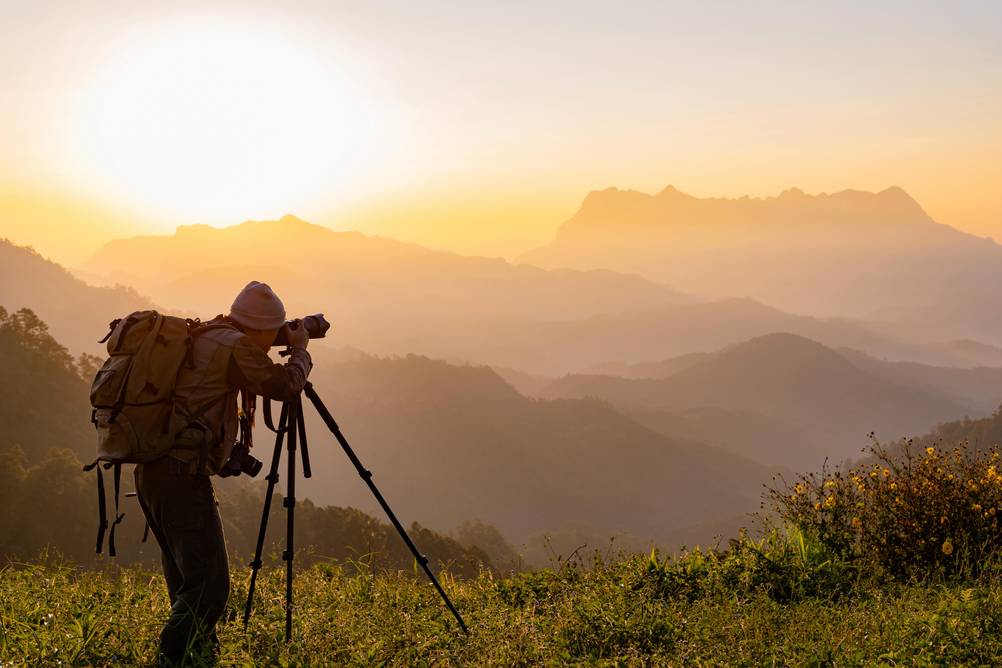 Reisestativ im Test: Eine Person steht bei Sonnenaufgang mit Kamera und Stativ vor einer Gebirgskulisse.