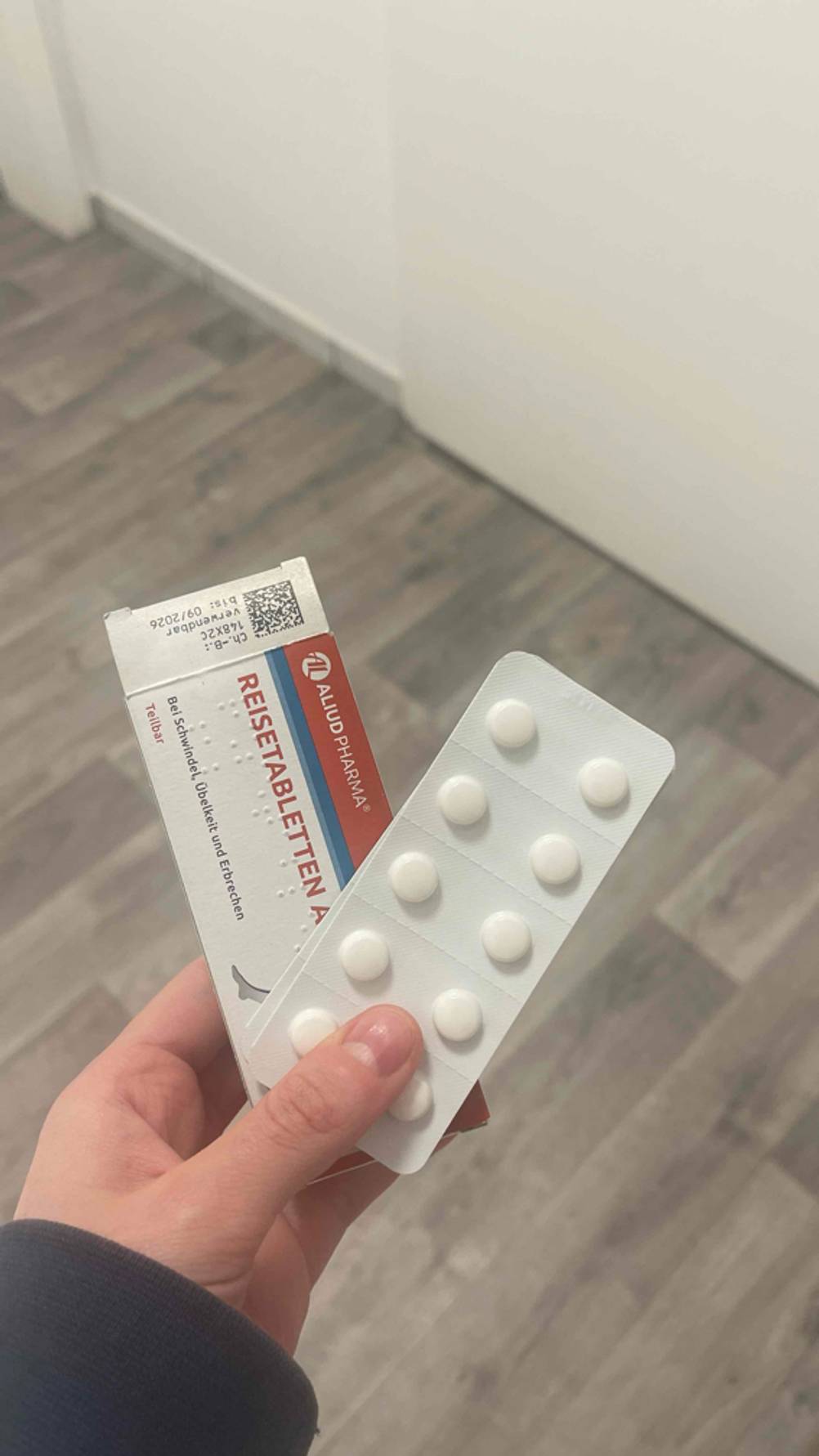 Eine Hand hält eine weiß-rot-blaue Verpackung und ein Blister mit 10 Tabletten über einem grauen Fußboden.
