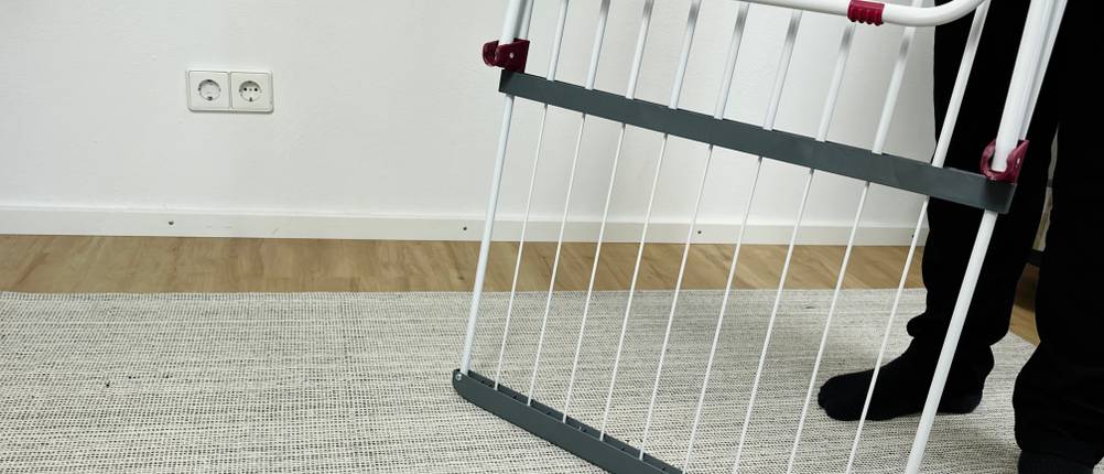 Der Relaxdays 10018866 steht seitlich zusammengeklappt auf dem Teppich. Eine Person hält den Wäscheständer an einer Seite fest.