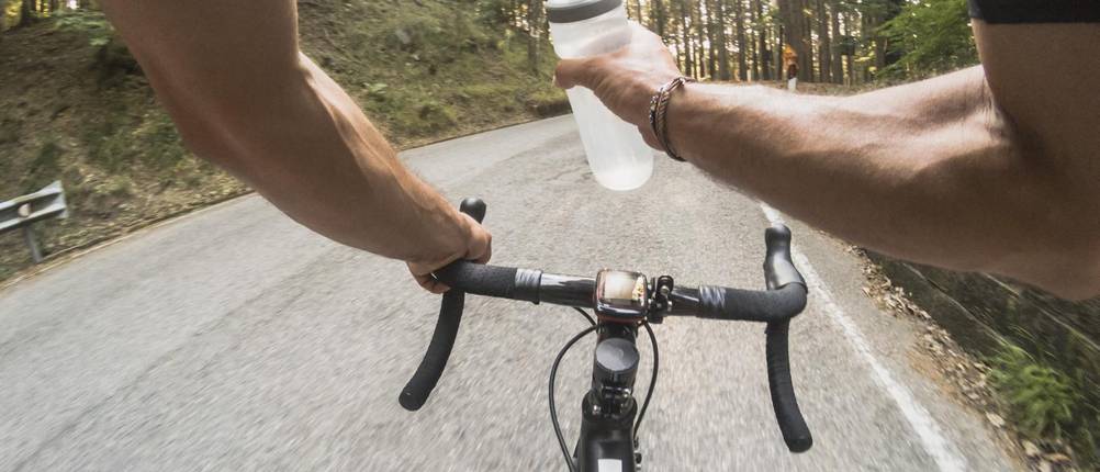 Rennrad-Flaschenhalter-Test: Ein Rennradfahrer fährt auf der Straße. Man sieht den Lenker. In einer Hand hält er eine Trinkflasche.