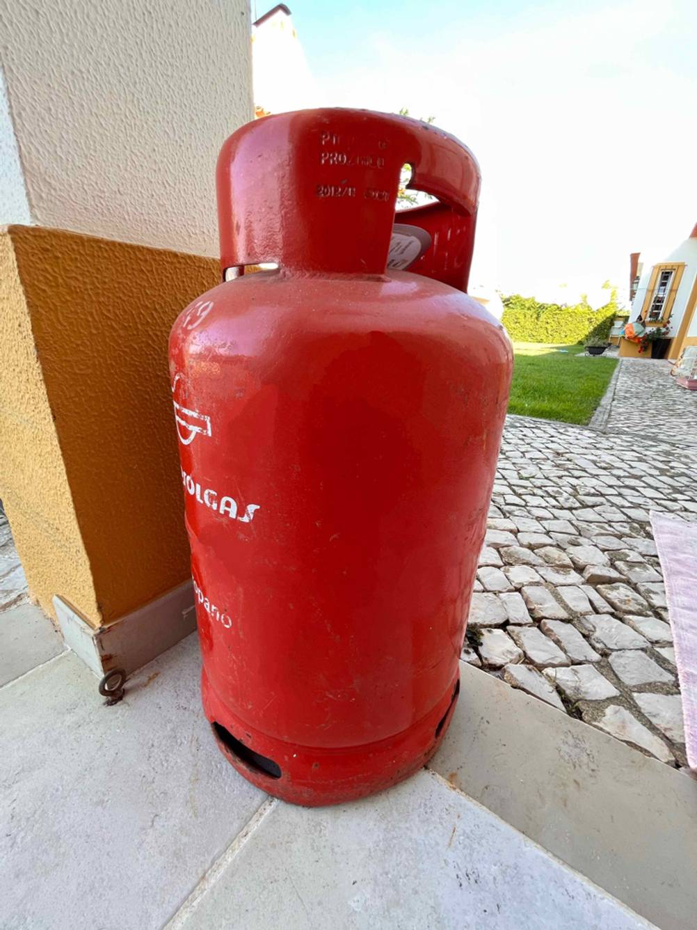 Eine rote Propangasflasche im Test steht an einem Pfeiler auf einer Terrasse im Gartenbereich.