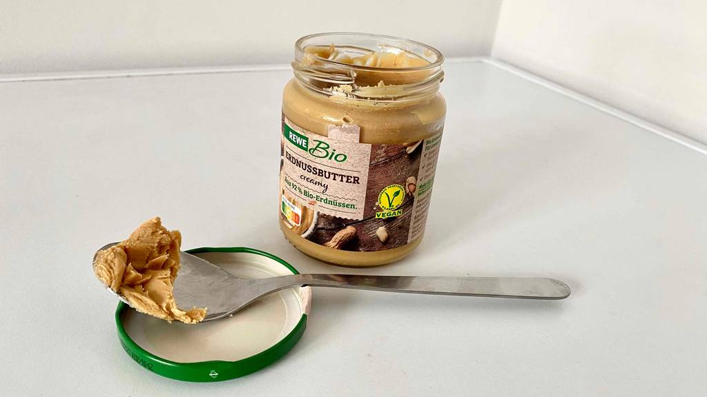 Ein Löffel mit getesteter Erdnussbutter liegt auf der Innenseite des Deckels vor einem geöffneten Glas der Marke Rewe.