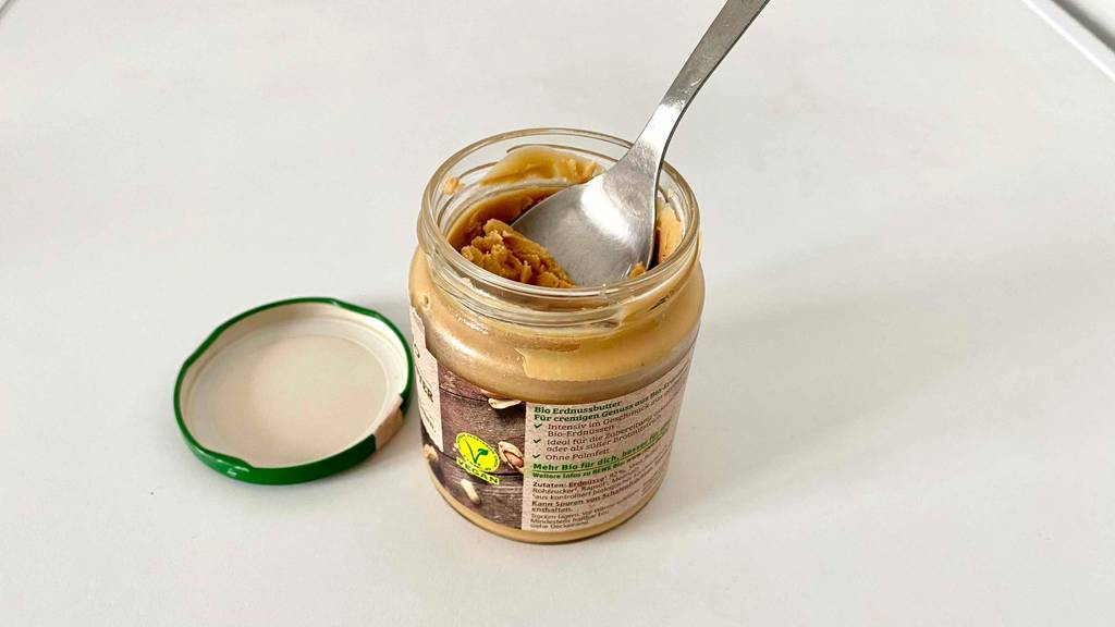Getestete Erdnussbutter im geöffneten Glas, in der ein Löffel steckt, befindet sich auf einer weißen Oberfläche.