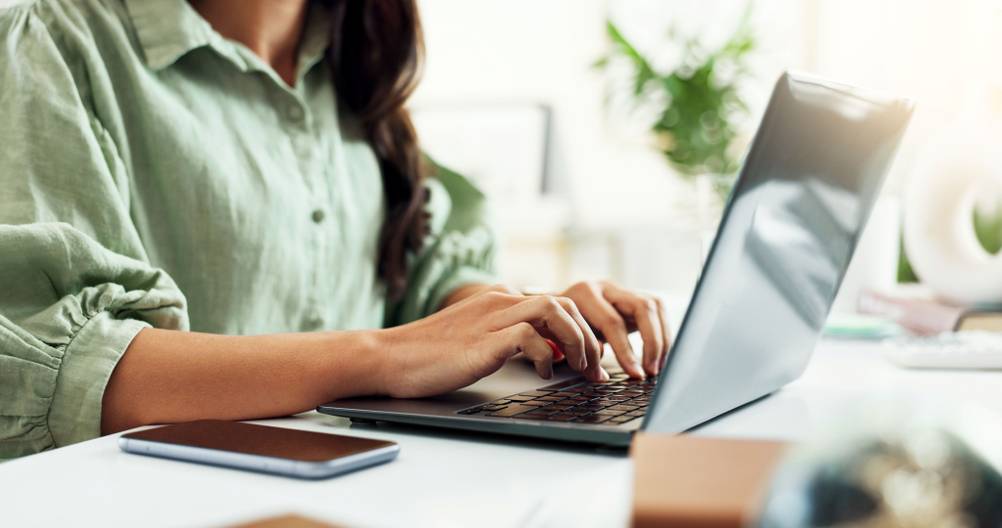 Eine Frau sitzt an einem weißen Schreibtisch und tippt auf der Tastatur eines Laptops.