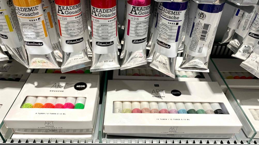 Gouachefarben im Test: mehrere weiße Verpackungen mit Tuben in Neon oder Pastellfarbe befinden sich in einem Verkaufsregal.
