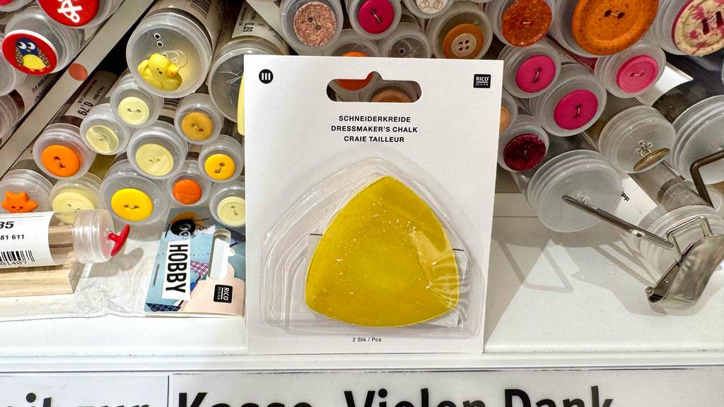 Test: Die Schneiderkreide in Gelb lehnt in der transparenten Verpackung vor einem Knopfregal im Geschäft.