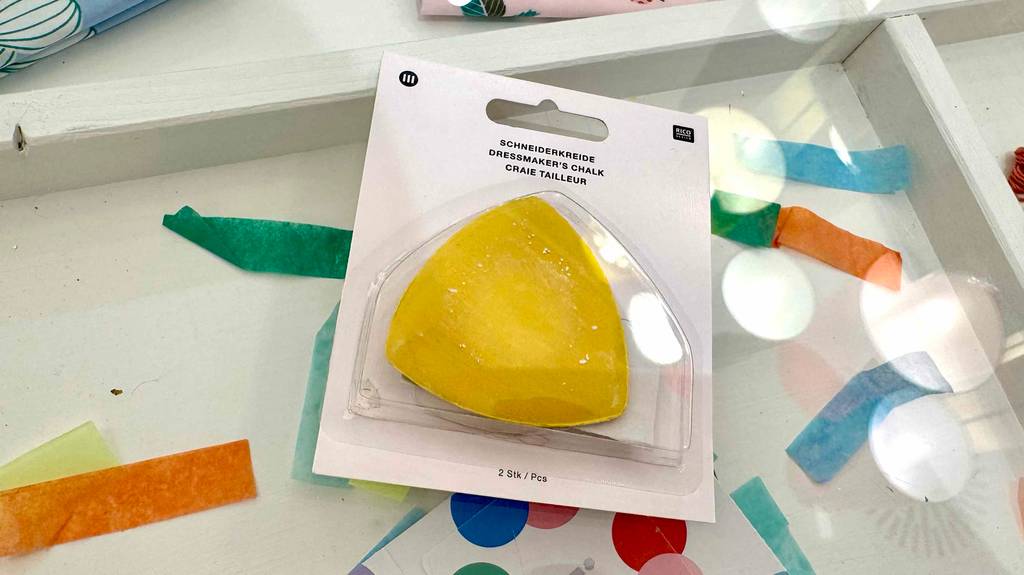 Die dreieckige getestete Schneiderkreide in Gelb liegt in zweifacher Ausführung innerhalb ihrer transparenten Verpackung auf bunten Papierfetzen.