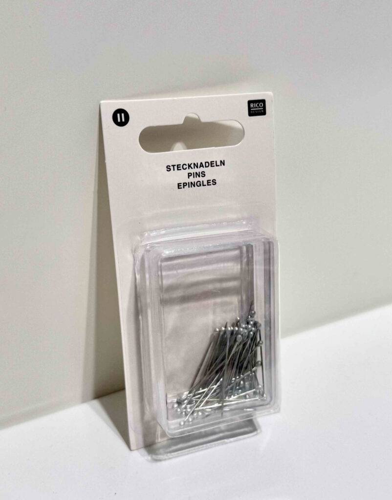 Eine transparente Packung mit Metall-Stecknadeln im Test lehnt an einer weißen Wand. 