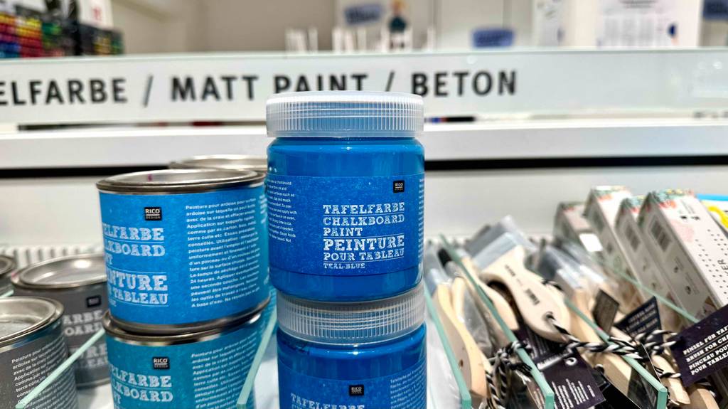 Blaue getestete Tafelfarben in Kunststoffdosen stehen übereinander neben Farbdosen auf einem Regal.
