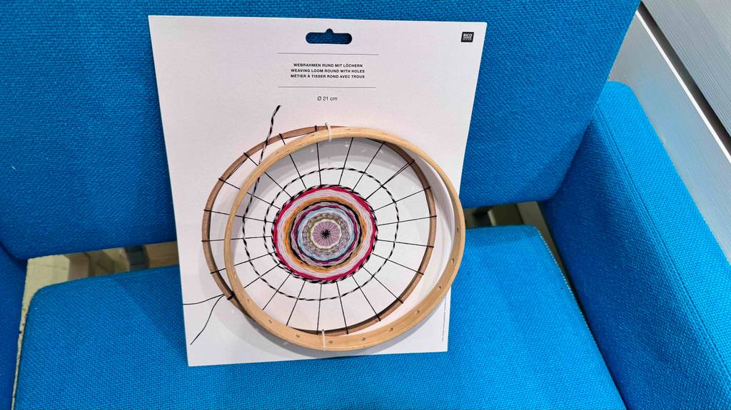 Der runde Holz-Webrahmen im Test steht inklusive seiner Kartonage-Verpackung auf einem blauen Sessel.