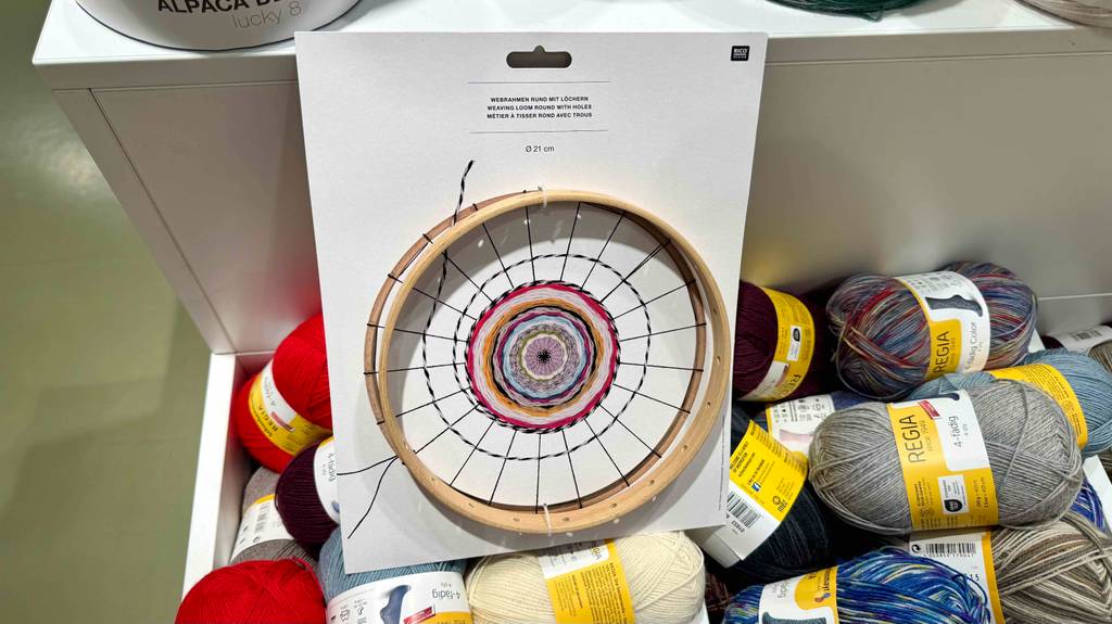 Der getestete Webrahmen befindet sich auf seiner Verkaufskartonage auf einem Haufen bunter Wollknäule im Geschäft.