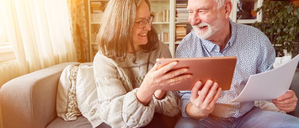 Riester-Rente im Test: Ein glückliches Ehepaar sitzt auf dem Sofa und schaut auf ein Tablet.