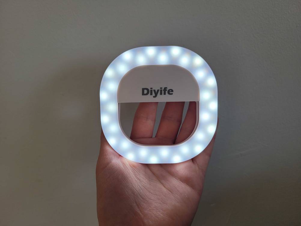 Eine Hand hält ein Ringlicht für das Handy im Test mit angeschaltetem blauen Licht vor eine helle Wand.