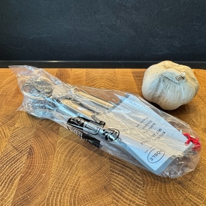 Die Rösle Knoblauchpresse in ihrer Plastikverpackung liegt neben einer Knoblauchknolle.