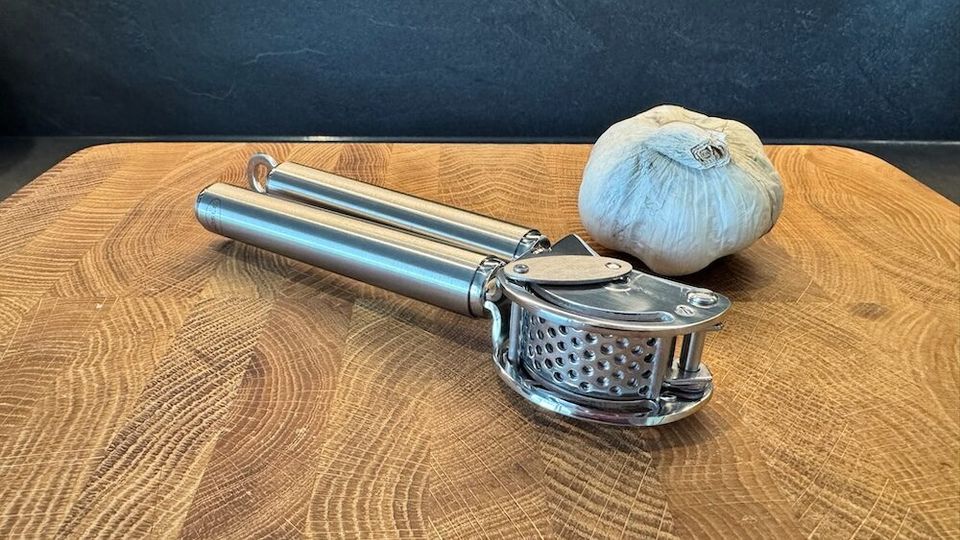 Eine Knoblauchpresse mit Abstreifer liegt neben einer Knoblauchknolle.