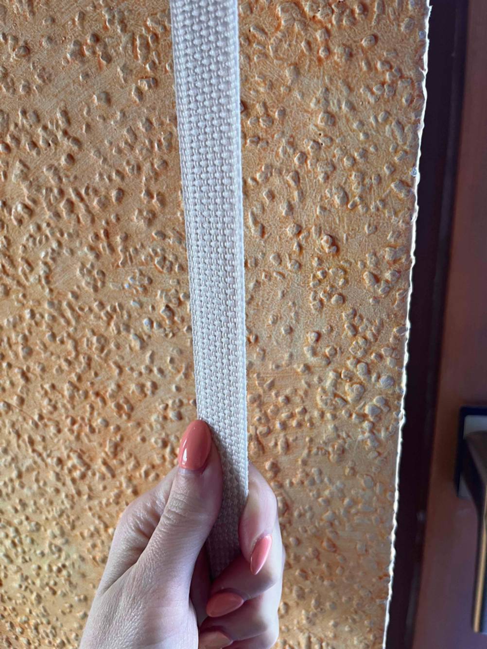 Eine Hand zieht einen weißen, getesteten Rollladengurt, der sich vor einer orangefarbenen Raufasertapete befindet.