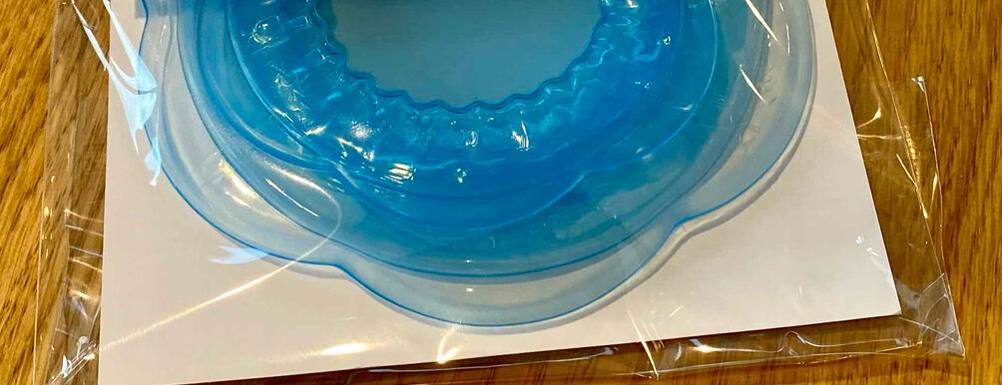 Detailansicht von zwei blau-transparenten Silikondeckeln im Test, die in einer Kunststoffverpackung auf einer Holzoberfläche liegen.