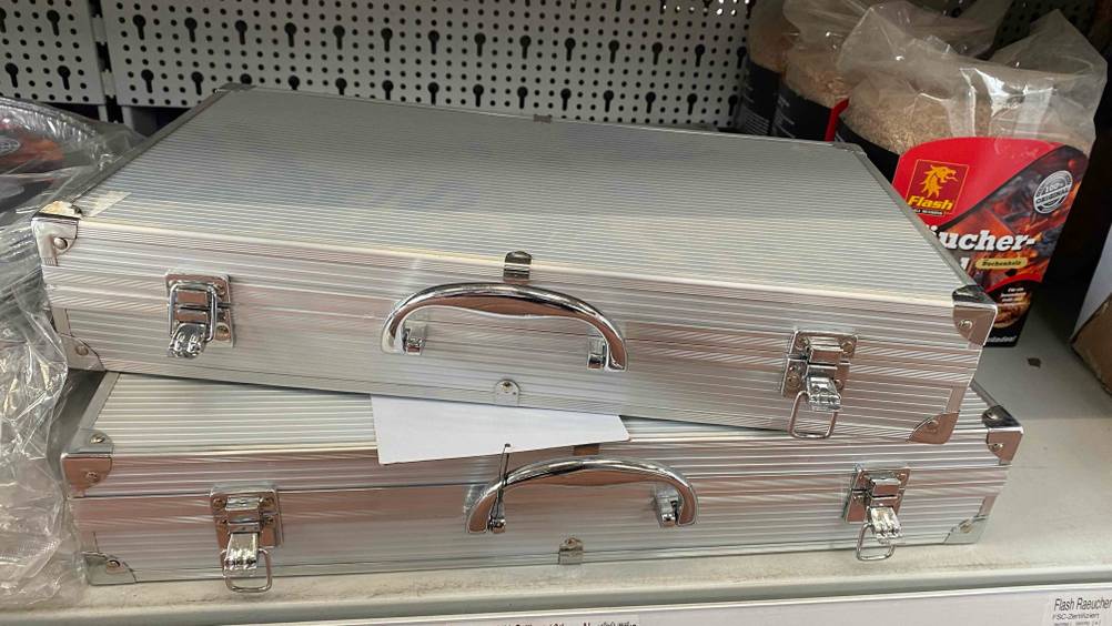 Im Test: Zwei zugeklappte Grillkoffer übereinander gestapelt in einem Verkaufsregal neben anderen Produkten.