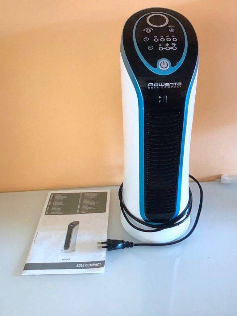Ein weiß-blau-schwarzer, getesteter leiser Ventilator von Rowenta steht neben seiner Bedienungsanleitung auf einem Glastisch vor oranger Wand.