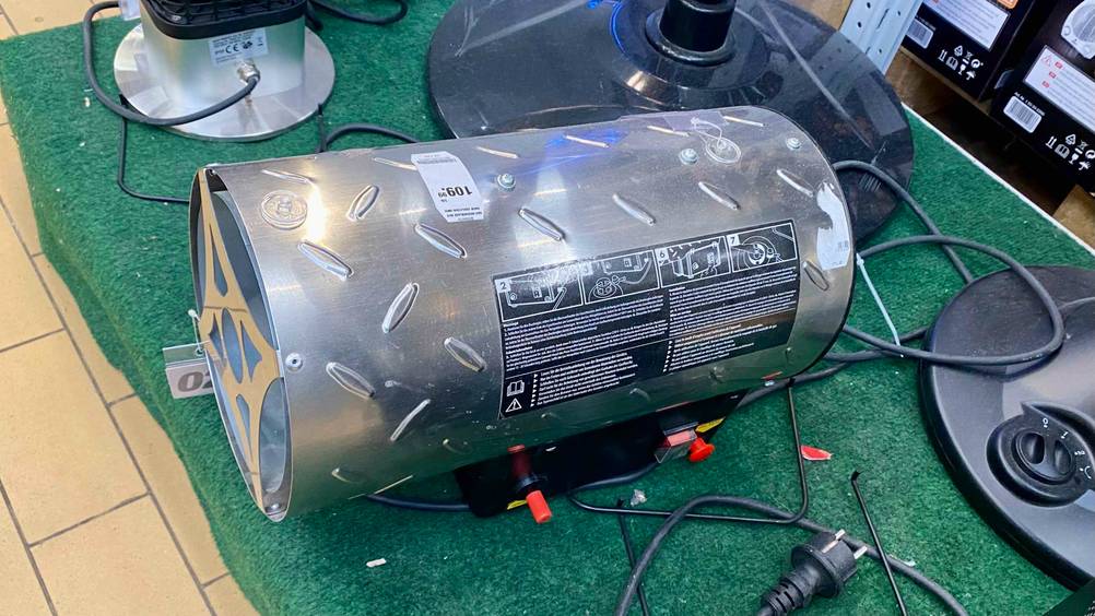 Ansicht von schräg oben auf einen auf grünem Kunstrasen stehenden Gasheizstrahler im Test, dessen Metall-Zylinder auf einer Elektro-Basis inklusive Bedienpanel mit roten Knöpfen montiert ist.
