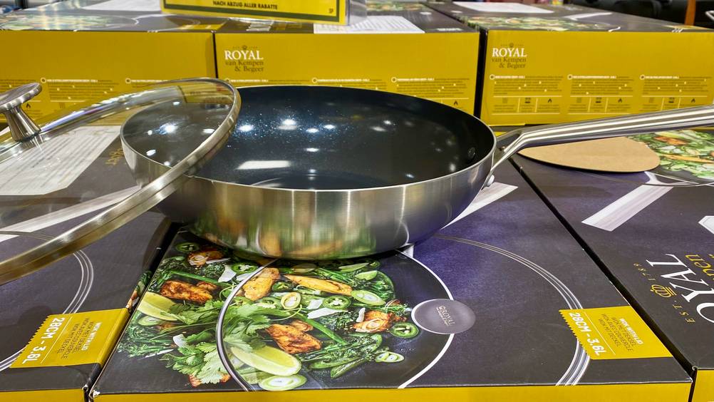 Wok mit Deckel im Test: Seitliche Ansicht einer Wokpfanne mit Fokus auf die Höhe des Randes steht inklusive angelehntem Glas-Deckel auf Originalverpackungen der Marke Royal.