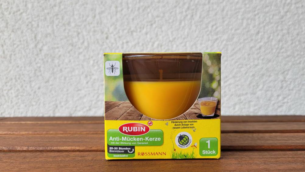 Eine originalverpackte Anti-Mücken-Kerze im Test mit Sichtfenster in der Packung auf einem Holztisch vor einer hellen Wand.