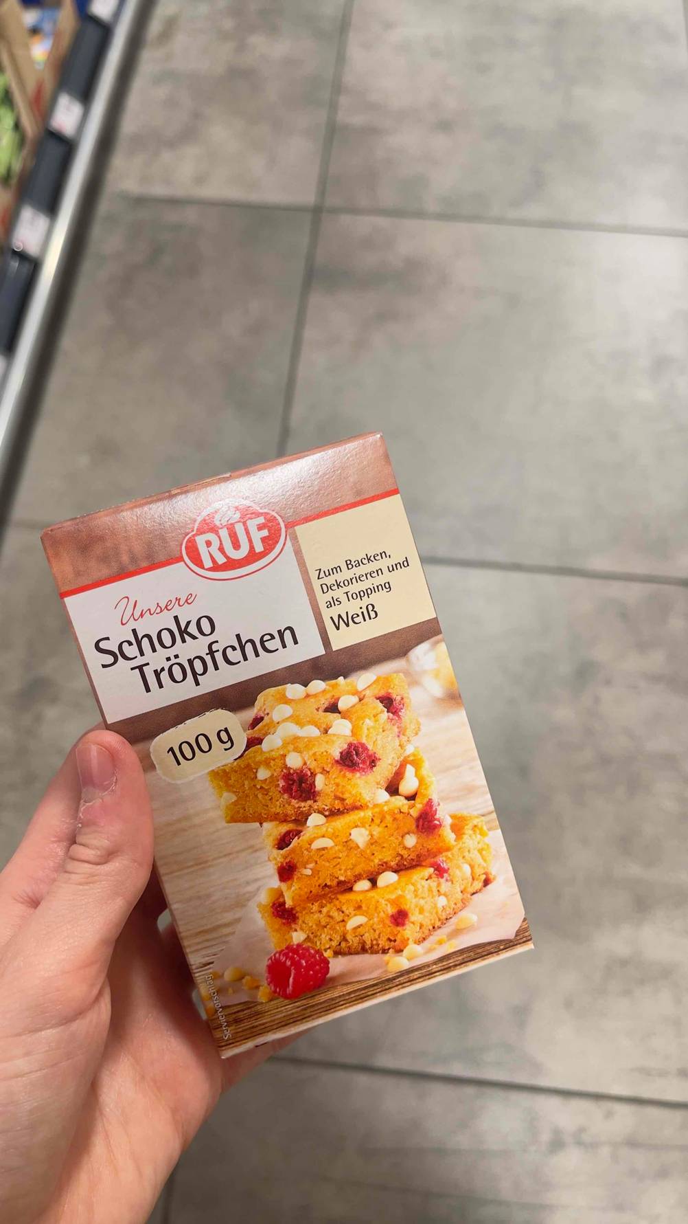 Im Test: Schokodrops der Marke RUF in der 100-g-Packung in einer Hand über einem grauen Fliesenboden.