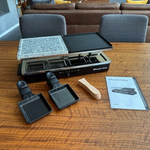 Das Russell Hobbs 26280-56 steht neben zwei Pfannen, den Spachteln und der Anleitung auf einem Tisch.