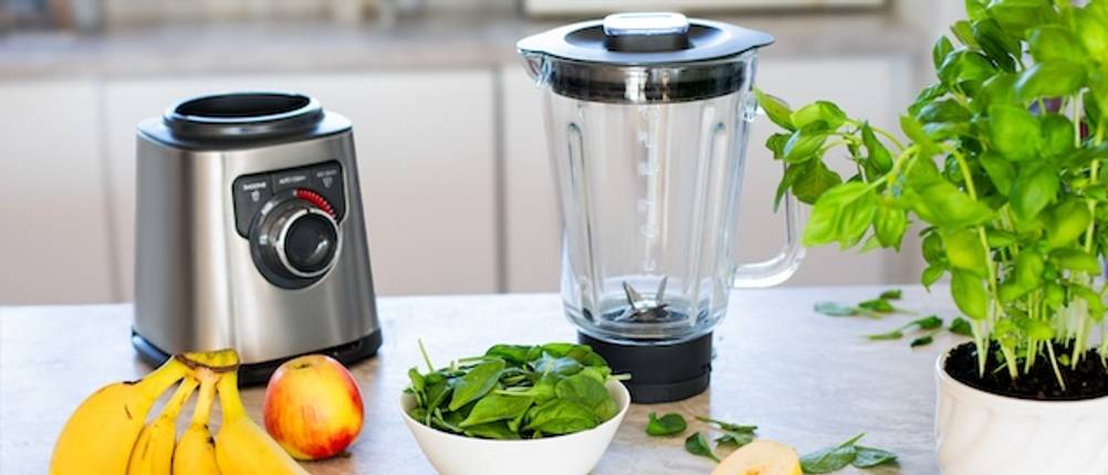 Russell-Hobbs-Standmixer im Funktions-Test: Das Gehäuse eines Standmixers aus Edelstahl und der Mixbehälter dazu stehen nebeneinander auf einer weißen Ablagefläche in einer Küche. Des weiteren befinden sich noch eine weiße Schüssel mit grünen Blättern sowie Obst auf der Ablage.