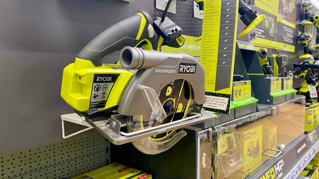 Eine bürstenlose gelb-graue Ryobi Kreissäge im Test, mit Fokus auf den Staubabsaug-Anschluss an einem grauen Ausstellungsregal neben weiteren Ryobi Produkten im Verkauf.