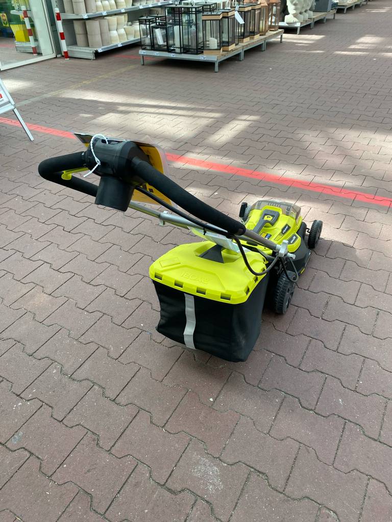 Gesamtansicht von hinten auf den gelben, getesteten Rasenmäher mit Mulchfunktion der Marke Ryobi im Außenbereich eines Gartencenters, inklusive schwarzem Fangkorb und höhenverstellbarem Griff.