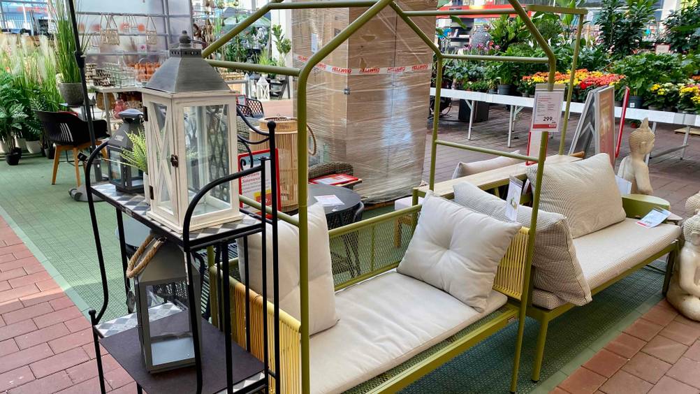 Gesamtansicht einer grün-gelb-orangen Gartenbank aus Metall im Test, die über ein Baldachin-Gestell verfügt und mit weißen Sitzkissen ausgestattet in einem Gartencenter steht.