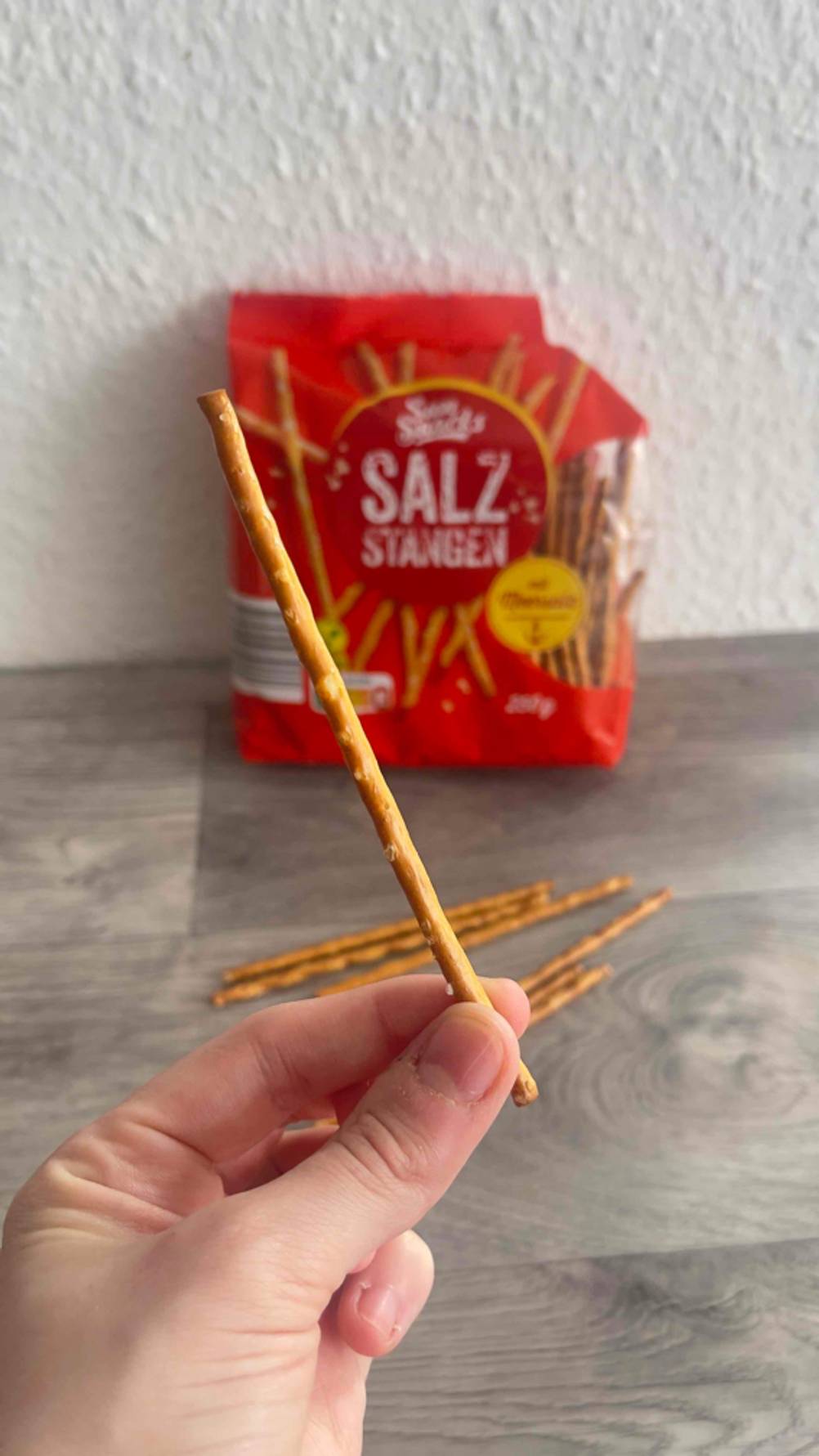 Eine Hand hält eine Salzstange zum testen zwischen den Fingern, im Hintergrund die Packung Salzstangen.
