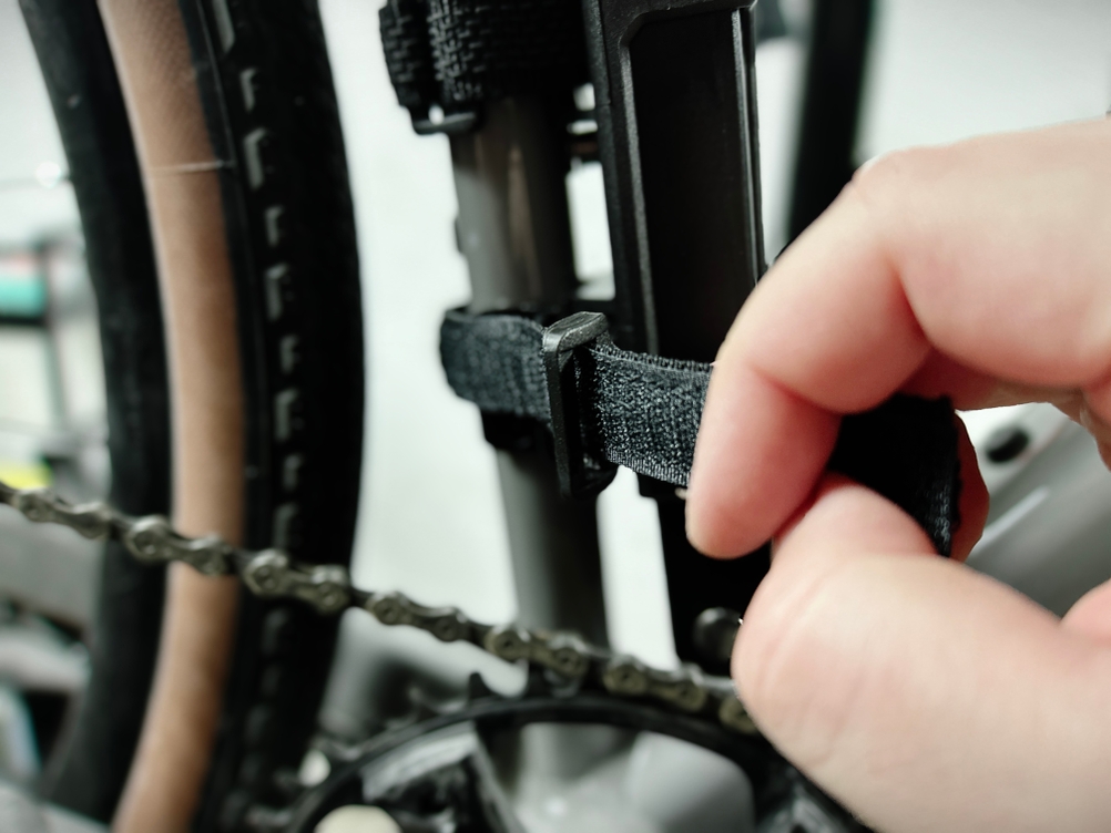 Eine Hand befestigt das Samok-Klappradschloss mit einem Klettband am Fahrradrahmen. Das Band wird durch die Halterung gezogen.