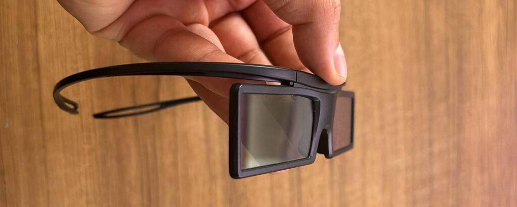 Von der Seite betrachtet hält eine Hand die 3D-Brille im Test vor einem Holzhintergrund.