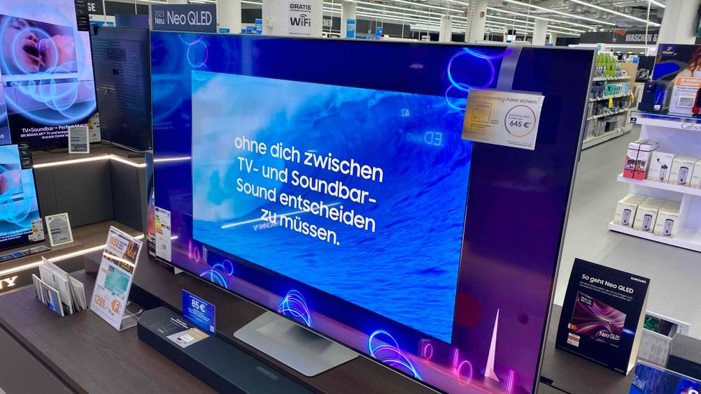 Ein getesteter Samsung-Fernseher (65 Zoll) in einer Verkaufsfläche mit Preisangaben auf einem braunen Sideboard.