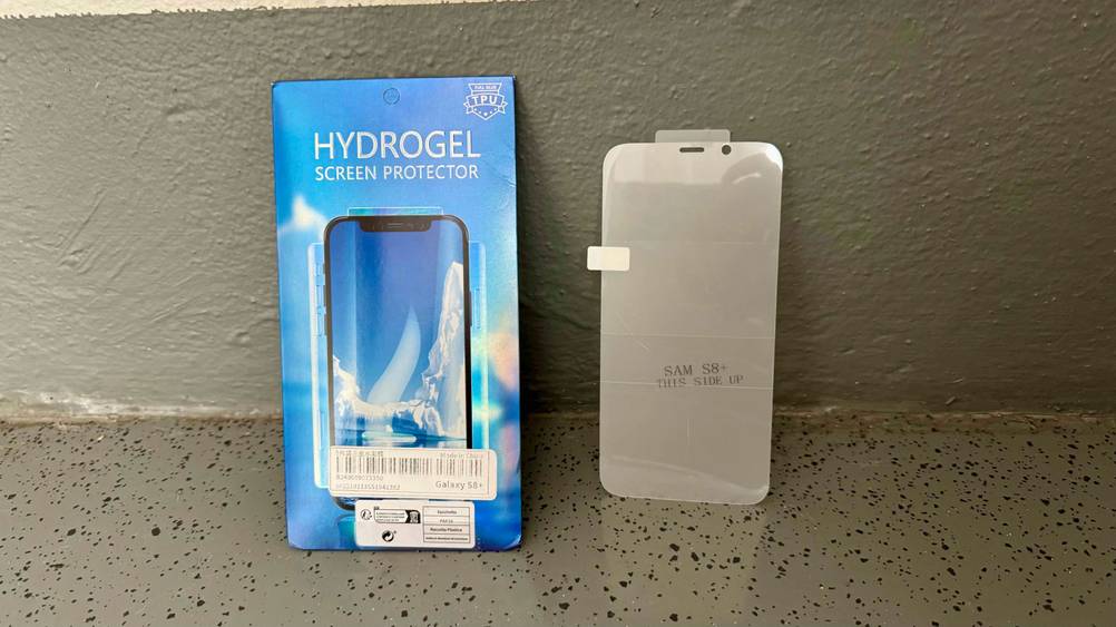 Neben der Originalverpackung steht ein getestetes Samsung-Galaxy S8-Plus-Panzerglas vor einer grauen Wand.