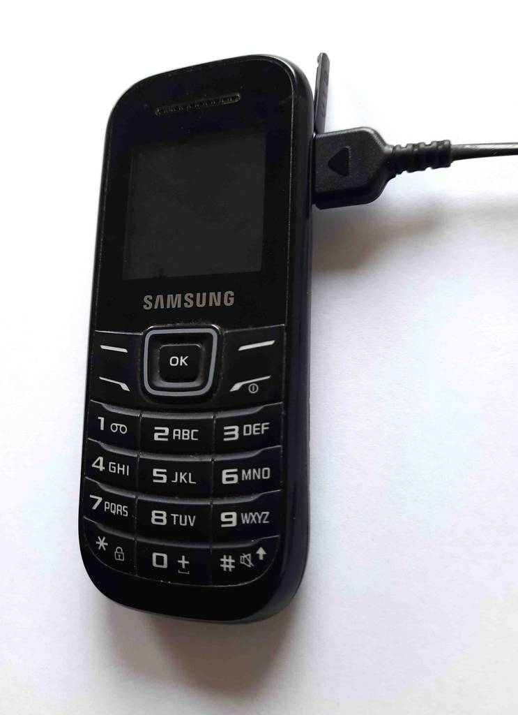 Getestet: Ein Tastenhandy in Schwarz von Samsung liegt inklusive eingestecktem USB-Anschlusskabel auf einem weißen Untergrund.