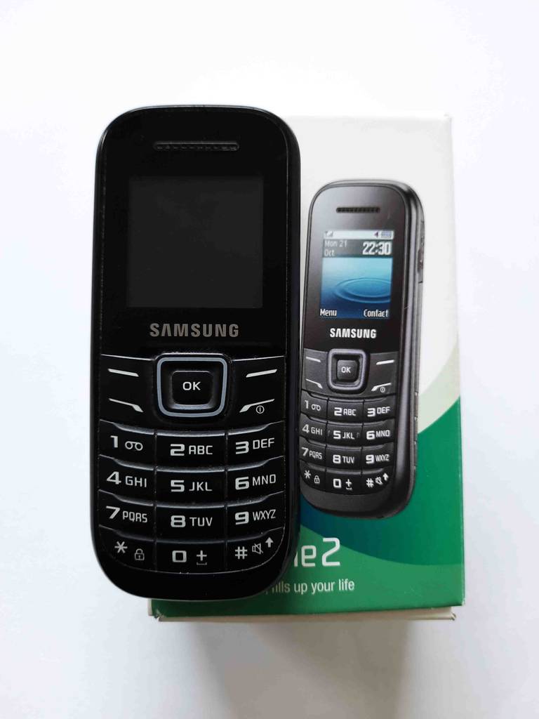 Im Test: ein Tastenhandy in Schwarz der Marke Samsung befindet sich vor seiner weiß-grünen Originalverpackung vor einem weißen Hintergrund.