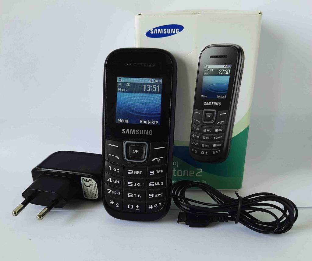 Ein getestetes Tastenhandy der Marke Samsung steht neben seinem Adapter und Kabel vor der Verpackung.