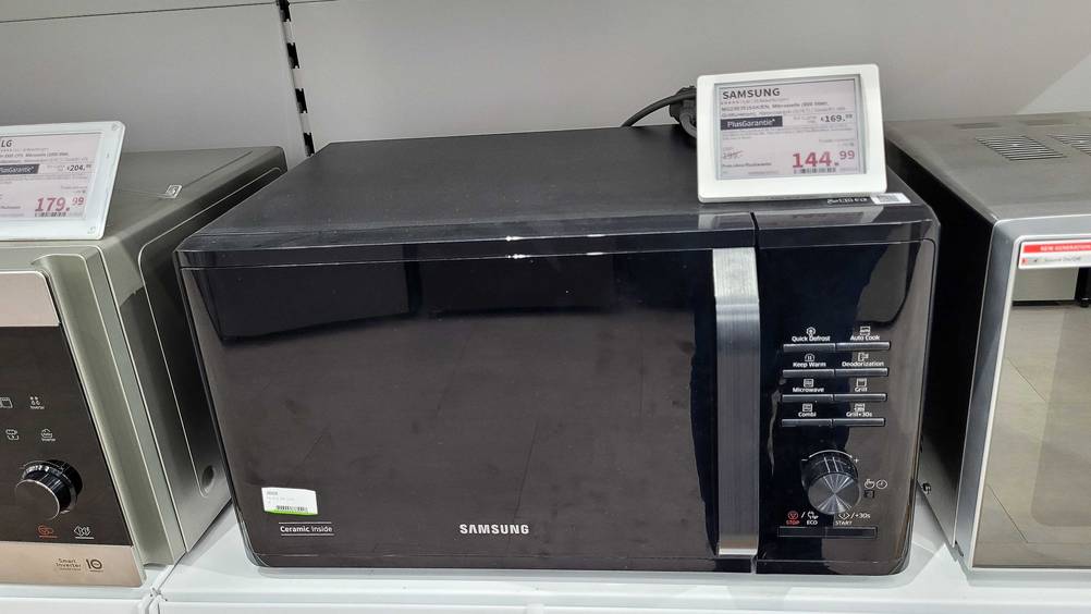 Eine getestete Samsung-Mikrowelle zwischen anderen Geräten in einem Regal mit Preisangabe.